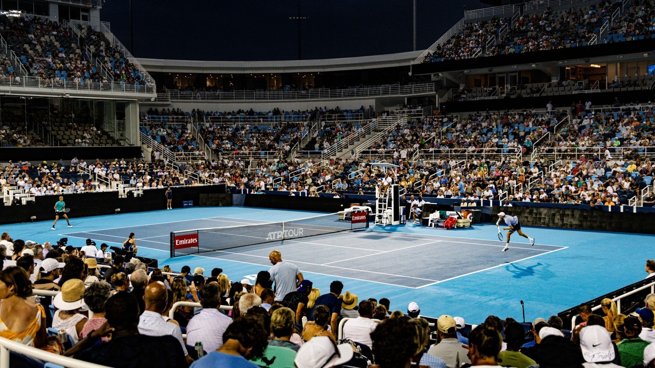 A full tennis stadium surrounds a blue hard court
